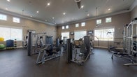 This fitness center is designed to accommodate a wide range of exercise needs within a single, open floor plan. The space features soaring ceilings accented by crown molding and recessed lighting, creating a bright and airy atmosphere. Clerestory windows positioned along the upper walls, combined with larger lower windows fitted with blinds, allow ample natural light to fill the room while maintaining privacy. Ceiling fans provide additional airflow throughout the space.

The flooring consists of durable rubber gym matting, which provides both cushioning and traction underfoot. The strength training area is well-stocked with multiple selectorized cable and resistance machines, including chest press, leg extension, and functional training stations. Large wall-mounted mirrors line one side of the room, visually expanding the space and allowing users to monitor their form during exercise.

The cardio section features Matrix-brand treadmills, elliptical trainers, upright stationary bikes, and a stair climber. Wall-mounted flat-screen televisions are positioned throughout for entertainment during workouts. Additional amenities include a weight rack with barbells, an adjustable bench, and a small storage unit for personal items near the entrance.