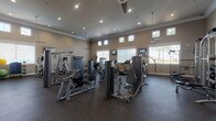 This fitness center is designed to accommodate a wide range of exercise needs within a single, open floor plan. The space features soaring ceilings accented by crown molding and recessed lighting, creating a bright and airy atmosphere. Clerestory windows positioned along the upper walls, combined with larger lower windows fitted with blinds, allow ample natural light to fill the room while maintaining privacy. Ceiling fans provide additional airflow throughout the space.

The flooring consists of durable rubber gym matting, which provides both cushioning and traction underfoot. The strength training area is well-stocked with multiple selectorized cable and resistance machines, including chest press, leg extension, and functional training stations. Large wall-mounted mirrors line one side of the room, visually expanding the space and allowing users to monitor their form during exercise.

The cardio section features Matrix-brand treadmills, elliptical trainers, upright stationary bikes, and a stair climber. Wall-mounted flat-screen televisions are positioned throughout for entertainment during workouts. Additional amenities include a weight rack with barbells, an adjustable bench, and a small storage unit for personal items near the entrance.