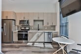 This single-floor unit in Atlanta, GA, offers a thoughtfully designed open layout that maximizes every inch of available space. The kitchen is a standout feature, showcasing light wood cabinetry with matte black hardware, sage green subway tile backsplash, white quartz countertops, and a full suite of stainless steel appliances including a gas range, microwave, refrigerator, and dishwasher. Natural light streams through large windows adjacent to the dining area, where a compact round table and chairs provide a functional eating space beneath a wall-mounted television.

The living and sleeping area is generously proportioned, benefiting from soaring ceilings, large floor-to-ceiling windows, and a ceiling fan. Warm wood-look flooring runs throughout, grounding the bright, neutral palette. A cozy sitting area complements the sleeping space, creating a versatile and inviting environment.

The bathroom is finished with a light wood vanity, bold black-framed mirror, white countertop, wood-look tile flooring, and a combined tub and tiled shower. A full-length mirror near the entryway adds depth to the corridor. The overall aesthetic is clean, contemporary, and well-appointed.