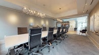 This professional conference space is thoughtfully designed to inspire productivity and impress guests. At its center sits a generously sized white conference table surrounded by high-back black mesh executive chairs with chrome frames, comfortably accommodating a large group. Overhead, a striking geometric chandelier with exposed bulb clusters serves as the room's focal point, complemented by a second pendant light fixture that casts a warm, inviting glow throughout the space. The flooring features a herringbone-patterned carpet in soft grey tones, adding texture and visual interest underfoot. One wall is adorned with a curated series of large framed abstract artworks in neutral whites and bold navy blue accents, illuminated by a dedicated track lighting system that highlights the pieces gallery-style. A credenza with mid-century modern styling and a projector provides functional presentation support, while a console table with decorative ceramic vessels adds an elegant finishing touch. Floor-to-ceiling glass partitions along one side of the room offer transparency and connectivity to adjacent spaces, with views hinting at a broader office environment beyond. Soft grey walls and white trim unify the refined, contemporary aesthetic throughout, creating a space that balances professionalism with sophisticated design.