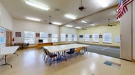 Located at 3500 E McKinney St in Denton, TX, this single-story community facility offers a generous and well-organized layout across a substantial floor plan. The space is designed to accommodate a variety of activities and gatherings, with multiple distinct areas connected by carpeted hallways adorned with warm wood trim, wainscoting, and a decorative transom window that invites natural light.

The large main activity hall is a standout feature, boasting soaring ceilings, ceiling fans, and rows of windows fitted with blinds that bathe the room in natural light. The hall includes a marked activity area on the carpet, flexible table-and-chair seating, and wall-mounted displays, making it well-suited for group events and recreational programs.

A second open room serves as an office and multi-purpose space, featuring high ceilings, colorful tile flooring, a workstation, bulletin boards, and French doors leading to adjacent areas. The cozy living room provides a relaxed lounge setting with upholstered seating, built-in wood cabinetry, and a television, all complemented by tall ceilings and French door access.

The fully equipped kitchen features warm amber walls, extensive oak cabinetry, hardwood flooring, a full suite of appliances, and ample counter space. Throughout the facility, consistent wood trim detailing, crown molding, and well-placed lighting contribute to a cohesive, welcoming ambiance.