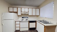 Located at 10714 Abercorn Ext in Savannah, Georgia, this single-floor unit offers a straightforward and functional layout throughout. The kitchen is well-equipped, featuring white cabinetry with wood-tone trim, a white electric range, dishwasher, refrigerator, and a stainless steel sink positioned beneath a window. Ample upper and lower cabinet storage lines the walls in an L-shaped configuration, and overhead fluorescent lighting ensures a bright workspace. Exposed ductwork and a textured ceiling add to the practical character of the room, while a secondary door provides convenient exterior access directly from the kitchen. The living area is open and neutral in tone, with warm beige walls, crisp white baseboards, and dark hardwood-style flooring that carries throughout the unit, lending a cohesive and clean aesthetic. A window allows natural light to fill the space, and multiple electrical outlets are thoughtfully placed along the walls. The bathroom includes a tiled tub-shower combination with a window above for ventilation and natural light, a toilet, a vanity with a medicine cabinet mirror, and a textured wall finish. The consistent use of dark flooring, neutral wall tones, and textured ceilings creates a uniform and tidy atmosphere throughout this practical Savannah residence.