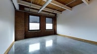 This single-floor space showcases a compelling industrial aesthetic that seamlessly blends raw architectural elements with a clean, open layout. The most striking features are the exposed red brick walls, which lend warmth and character to the interior, complemented by polished concrete floors that reflect natural light and add a sleek, modern contrast. The ceiling is finished with warm-toned wooden planks supported by structural beams, while exposed metal ductwork and piping run overhead, reinforcing the space's industrial character. Large black-framed windows along the brick wall allow natural light to pour in, brightening the interior and providing views to the outside. The open-plan design offers exceptional flexibility, making the space well-suited for a variety of uses, including creative studios, retail, office environments, or event venues. Light-colored walls on the opposing side of the room provide a neutral backdrop that balances the richness of the brick and wood tones. The overall ambiance is one of urban authenticity — a thoughtfully preserved industrial environment that offers both character and adaptability for a wide range of occupants and purposes.