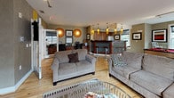 Located at 400 Blake St in New Haven, CT, this single-floor community lounge is a well-appointed gathering space designed for both relaxation and recreation. The open-plan layout is anchored by warm hardwood flooring that flows seamlessly throughout the entire area, creating a cohesive and inviting atmosphere.

The main lounge area features multiple plush sofas arranged around a glass coffee table, providing comfortable seating for socializing. A wall-mounted flat-screen TV, track lighting overhead, and large windows that invite ample natural light and views of the surrounding grounds complete the living area. Decorative framed artwork and globe-style wall accents add character to the textured, neutral-toned walls.

A striking pool table with a bold red felt surface serves as a central recreational focal point, positioned in a generously open section of the room. Glass doors lead directly to an outdoor area, further enhancing the sense of space and accessibility.

The kitchen is thoughtfully designed with rich wood cabinetry, dark granite countertops, and a dramatic speckled black tile backsplash. Stainless steel appliances, including a dishwasher and microwave, offer full functionality. Warm pendant lighting in amber tones hangs above a tiled breakfast bar with bar stool seating, creating a distinct yet connected dining area. A workstation area is also visible, adding a practical dimension to this multifunctional community space.