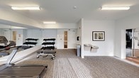 This fitness center is thoughtfully designed to support a wide range of workout routines across two distinct zones. The main exercise area features durable carpet flooring and is outfitted with cardio machines including treadmills and an elliptical, along with a multi-tiered dumbbell rack and adjustable weight benches. Full-length mirrored walls line one side of the room, visually expanding the space and allowing users to monitor their form during workouts. A wall-mounted flat-screen television adds convenience for those looking to stay entertained during cardio sessions. Hydration stations, including a dual-level water fountain, are conveniently positioned along the wall, alongside a paper towel dispenser for equipment maintenance. Bright fluorescent lighting ensures the space is well-illuminated throughout. Adjacent to the main area, a separate studio room features warm hardwood flooring, a bold blue accent wall, and large sliding glass doors that invite natural light. This studio is equipped with cycling bikes, a ballet barre with mirrored wall, kettlebells, medicine balls, and a foam roller, making it well-suited for group fitness classes, cycling sessions, or individual training. A barn-style sliding door connects the two areas seamlessly. The overall ambiance is clean, modern, and functional.