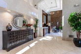 Located at 13 Vose Ave in South Orange, NJ, this single-floor lobby space serves as the welcoming gateway to The Avenue at South Orange. The entrance is defined by a set of floor-to-ceiling glass doors framed in dark metal, flooding the interior with natural light and providing clear sightlines to the surrounding streetscape. A branded entry mat bearing the property's name greets visitors upon arrival.

The lobby's soaring double-height ceilings create an impressive sense of volume and openness. Polished large-format tile flooring runs throughout, reflecting natural light and contributing to the space's bright, refined atmosphere. The reception area features a rich, dark wood desk paired with an elegant marble-topped counter, accented by a textured mosaic feature wall in warm golden tones that serves as a striking focal point.

A comfortable seating arrangement with sleek, contemporary black chairs and a glass-topped coffee table provides a welcoming lounge area. Lush potted plants are strategically positioned throughout, adding organic warmth to the modern design. Decorative chandelier lighting complements the recessed ceiling fixtures. A staircase and elevator are accessible from the lobby, and an arched architectural detail adds a classic touch to the otherwise contemporary interior.