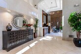 Located at 13 Vose Ave in South Orange, NJ, this single-floor lobby space serves as the welcoming gateway to The Avenue at South Orange. The entrance is defined by a set of floor-to-ceiling glass doors framed in dark metal, flooding the interior with natural light and providing clear sightlines to the surrounding streetscape. A branded entry mat bearing the property's name greets visitors upon arrival.

The lobby's soaring double-height ceilings create an impressive sense of volume and openness. Polished large-format tile flooring runs throughout, reflecting natural light and contributing to the space's bright, refined atmosphere. The reception area features a rich, dark wood desk paired with an elegant marble-topped counter, accented by a textured mosaic feature wall in warm golden tones that serves as a striking focal point.

A comfortable seating arrangement with sleek, contemporary black chairs and a glass-topped coffee table provides a welcoming lounge area. Lush potted plants are strategically positioned throughout, adding organic warmth to the modern design. Decorative chandelier lighting complements the recessed ceiling fixtures. A staircase and elevator are accessible from the lobby, and an arched architectural detail adds a classic touch to the otherwise contemporary interior.