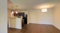 This single-floor residence offers a thoughtfully designed open-concept layout that seamlessly connects the kitchen and living areas. The rich, dark hardwood flooring runs throughout the space, providing a warm and cohesive foundation that complements the overall design aesthetic.

The kitchen is a standout feature, centered around a peninsula with granite countertops and an integrated sink, ideal for both meal preparation and casual dining. Dark espresso cabinetry provides ample storage and creates a striking contrast against the light-toned walls. A suite of black appliances, including a refrigerator, microwave, and range, adds a sleek, uniform finish to the kitchen. Stylish pendant lights hang above the peninsula, enhancing both functionality and visual appeal. A track lighting system further illuminates the space with directional flexibility.

The adjacent living area is open and spacious, bathed in warm ambient light from a large drum pendant fixture. The neutral wall tones provide a versatile backdrop, ready to accommodate a variety of furnishing styles. A paneled entry door anchors one end of the living space, offering a clean and classic architectural touch. The overall ambiance of the residence is modern yet inviting, with a well-balanced mix of dark and light tones creating a comfortable and stylish environment.