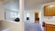 This single-floor unit offers a straightforward and functional layout designed to accommodate multiple occupants comfortably. The carpeted living room serves as the central gathering space, furnished with a sofa and armchairs, and benefits from natural light through a window with outdoor views. A textured ceiling adds a classic touch to the common areas, while baseboard heating provides warmth throughout.

The kitchen is equipped with warm oak cabinetry — both upper and lower — alongside a white electric range, refrigerator, and a single-basin sink with a light-colored countertop. Vinyl flooring in the kitchen provides easy maintenance, and there is space for a small dining table adjacent to the cooking area.

Each bedroom features plush carpeting, neutral white walls, and a window for natural light. The rooms are set up with a bed frame, mattress, and a desk with a chair, making them well-suited for student or shared living arrangements. Closets with hanging rods provide practical storage in the bedrooms.

The bathroom includes a full tub-shower combination, toilet, and vanity with wood-toned cabinetry, finished with vinyl flooring. A central hallway connects all areas of the unit efficiently, with wood-toned doors throughout maintaining a cohesive aesthetic.