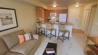 This single-floor unit is designed around an open-concept layout that seamlessly connects the kitchen and living area. The kitchen is outfitted with warm honey-toned wood cabinetry, a white refrigerator, over-the-range microwave, and a stovetop, providing ample functionality within a well-organized footprint. A peninsula counter with a light-toned surface extends outward, offering casual bar seating for three, making it an ideal spot for dining or entertaining. The countertop transitions smoothly into the surrounding space, reinforcing the open flow of the layout.

The living area features plush, light beige wall-to-wall carpeting that adds warmth underfoot, contrasting with the hard flooring visible near the entryway. A neutral-toned sofa accented with a plaid throw pillow anchors the sitting area, complemented by a dark rectangular coffee table. Walls are painted in a soft, warm beige, contributing to a cohesive and inviting ambiance throughout. Flush-mount ceiling lights provide consistent illumination. The entryway, visible in the background, features a white door and transitions cleanly into the main living space.