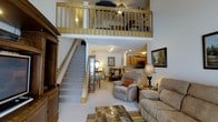 This two-story space showcases an open and airy design highlighted by soaring double-height ceilings in the main living area. The ground floor centers around a comfortable living room furnished with plush seating, a patterned area rug, and a large entertainment unit. Recessed and track lighting illuminate the space effectively throughout. A carpeted staircase with a natural wood railing leads to the upper loft level, which overlooks the main living area through a matching wood balustrade, adding architectural interest and a sense of openness. Adjacent to the living room, a dining area with traditional-style seating connects seamlessly to the kitchen, which features warm wood cabinetry, built-in appliances, and a breakfast bar. A glass-front display cabinet adds a decorative touch near the staircase. The consistent use of light-toned carpet, white walls, and natural wood accents throughout both levels creates a cohesive and welcoming ambiance. The loft space above provides additional functional square footage, visible from below and ideal for use as a bedroom or secondary living area.