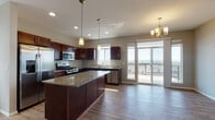 Located at 8808 North Indian Trail Road in Spokane, WA, this two-floor residence offers a well-appointed layout with modern finishes throughout. The open-concept main living area flows seamlessly from the entryway into the kitchen and dining space, all unified by light hardwood-style flooring. The kitchen is a standout feature, showcasing rich dark cabinetry, granite countertops, a large center island with pendant lighting, and a full suite of stainless steel appliances including a gas range, microwave, dishwasher, and French door refrigerator. Sliding glass doors open from the kitchen and dining area onto a private balcony, offering sweeping panoramic views of the surrounding landscape. A ceiling fan with lighting anchors the living area, adding both comfort and style. The bathrooms continue the design language with granite-topped dark wood vanities, hardwood-style flooring, and clean white fixtures, including a full tub and shower combination. Bedrooms are carpeted for warmth and comfort, with large windows that invite natural light and frame scenic outdoor views. A dedicated utility area with a sink and granite countertop provides added functionality. The overall ambiance is clean, modern, and move-in ready.