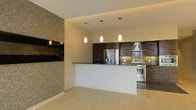 This single-floor space showcases a contemporary open-concept layout that seamlessly connects the living and kitchen areas. The kitchen is a standout feature, equipped with rich, dark wood cabinetry that provides ample storage and a striking visual contrast against the neutral-toned walls. Stainless steel appliances, including a French door refrigerator and a gas range with a stainless hood, lend a professional, polished feel to the culinary space. A peninsula with a dark countertop serves as both a functional workspace and a natural divider between the kitchen and the adjoining living area, making it ideal for casual dining or entertaining. Three elegant pendant lights hang above the peninsula, providing focused task lighting while adding a refined decorative touch. The kitchen also features a tasteful tile backsplash that complements the overall design. Light hardwood-style flooring runs throughout, brightening the space and enhancing its open feel. A textured accent wall adds depth and visual interest, while the clean white walls and recessed ceiling details contribute to the modern, cohesive aesthetic of the space.
