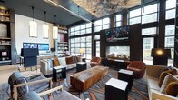 This stylish single-floor lounge space exudes a contemporary and refined atmosphere, thoughtfully designed for both comfort and visual impact. The room is anchored by a sleek, modern fireplace set beneath a large wall-mounted flat-screen television, creating a natural gathering focal point framed by rich, dark wall paneling. Generous floor-to-ceiling industrial-style windows flood the space with abundant natural light, offering views of the surrounding urban landscape and reinforcing a sense of openness and connectivity to the outside world. The ceiling is a striking artistic feature in its own right, adorned with a textured, mosaic-like treatment in muted earth tones that adds depth and visual intrigue overhead. A curated mix of furnishings — including plush neutral sofas accented with rust and amber throw pillows, supple leather swivel chairs, and a remarkable raw-edge wooden bench serving as a centerpiece coffee table — balances warmth with sophistication. Geometric side tables in black and white marble tones complement the boldly patterned area rug, which ties the space together with sweeping organic motifs. Along one wall, built-in shelving stocked with books and decorative objects adds an intellectual, lived-in character, while pendant lighting and recessed fixtures ensure a warm, ambient glow throughout.
