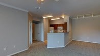 This single-floor residence features an open-concept design that seamlessly connects the living area to the kitchen. The living space is finished with neutral wall-to-wall carpeting and painted in a soft gray tone, creating a calm, understated ambiance throughout. Crown molding adds a refined architectural detail along the ceiling line, while a ceiling fan provides both function and a subtle design element overhead.

The kitchen is positioned toward the rear of the open space and is defined by a raised breakfast bar peninsula with a light stone-style countertop, offering a natural division between the kitchen and living area. Warm-toned wood cabinetry lines the kitchen walls, complemented by white appliances including a refrigerator and microwave. Overhead fluorescent lighting illuminates the kitchen workspace efficiently.

A hallway visible from the living area leads to additional rooms, with a set of double closet doors suggesting generous storage options. The overall layout is practical and cohesive, with consistent finishes and neutral tones that offer a versatile backdrop suitable for a variety of interior styles.