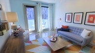 This single-floor space showcases a thoughtfully designed living area that balances modern aesthetics with a warm, inviting atmosphere. The room is anchored by a tufted gray sofa adorned with bold navy and coral accent pillows, complemented by a geometric multi-colored area rug in shades of orange, yellow, and red that adds a dynamic visual element to the polished floor beneath. A low-profile wooden coffee table serves as a functional centerpiece, styled with tasteful decorative objects.

Natural light floods the space through floor-to-ceiling glass doors framed by sheer white curtains, opening directly onto a private balcony furnished with outdoor seating. The balcony access enhances the sense of openness and connectivity to the outside. A sleek brass arc floor lamp provides additional ambient lighting, while framed wall art in red and black tones adds a curated, gallery-like quality to the white walls. The overall palette of soft blues, whites, and warm accent colors creates a cohesive, contemporary feel throughout the space, making it both visually engaging and functionally comfortable.