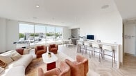 This sophisticated single-floor amenity space is defined by its bright, open layout and contemporary design. The interior is anchored by elegant herringbone-patterned light wood flooring that runs throughout, complementing the crisp white walls and recessed lighting overhead. Floor-to-ceiling windows and sliding glass doors flood the space with abundant natural light while offering sweeping panoramic city views, including glimpses of palm trees and the surrounding urban skyline.

The main lounge area features plush, terracotta-hued curved armchairs paired with a cream sectional sofa and a minimalist white coffee table, creating a warm and inviting gathering space. Adjacent to this seating zone, a foosball table positioned against the corner windows adds a recreational element. A wall-mounted flat-screen television, flanked by rich wood cabinetry and framed artwork, defines a dedicated media viewing area.

A business workstation lines one wall, offering a sleek counter with bar-height seating, desktop computers, and a printer — ideal for productivity. A bold chevron-patterned tile accent wall frames a small dining nook nearby, adding architectural interest. The space also includes an ADA-accessible restroom with clean, functional finishes. Access to an outdoor terrace further extends the space's versatility and connection to the city beyond.