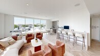 This sophisticated single-floor amenity space is defined by its bright, open layout and contemporary design. The interior is anchored by elegant herringbone-patterned light wood flooring that runs throughout, complementing the crisp white walls and recessed lighting overhead. Floor-to-ceiling windows and sliding glass doors flood the space with abundant natural light while offering sweeping panoramic city views, including glimpses of palm trees and the surrounding urban skyline.

The main lounge area features plush, terracotta-hued curved armchairs paired with a cream sectional sofa and a minimalist white coffee table, creating a warm and inviting gathering space. Adjacent to this seating zone, a foosball table positioned against the corner windows adds a recreational element. A wall-mounted flat-screen television, flanked by rich wood cabinetry and framed artwork, defines a dedicated media viewing area.

A business workstation lines one wall, offering a sleek counter with bar-height seating, desktop computers, and a printer — ideal for productivity. A bold chevron-patterned tile accent wall frames a small dining nook nearby, adding architectural interest. The space also includes an ADA-accessible restroom with clean, functional finishes. Access to an outdoor terrace further extends the space's versatility and connection to the city beyond.