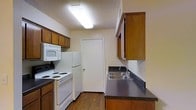 This single-floor residential space centers around a practical, well-equipped kitchen that makes efficient use of its layout. The kitchen features warm wood-toned upper and lower cabinetry that provides ample storage, complemented by dark laminate countertops and a double stainless steel sink. Essential appliances are included, among them a white electric coil-top range, an over-the-range microwave, and a full-size refrigerator, making the space move-in ready. A textured ceiling with a flush-mounted fluorescent light fixture ensures the kitchen is well-lit throughout. The wood-look flooring adds a touch of warmth to the space and runs through the length of the kitchen. A door at the far end of the kitchen leads to an additional area, offering convenient internal access. The overall layout of the kitchen is designed for functionality, with a galley-style configuration that places appliances and workspace within easy reach. The neutral wall tones and classic cabinetry create a timeless, practical aesthetic that is well-suited for everyday living.
