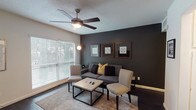 This single-story 2-bedroom, 1-bathroom unit offers a thoughtfully designed layout with dark hardwood-style flooring that flows throughout the space, creating a cohesive and modern aesthetic. The living room makes a bold statement with a dramatic dark accent wall, ceiling fan with light fixture, and large windows fitted with blinds that allow ample natural light to fill the room. The space comfortably accommodates a seating area centered around a modern coffee table, making it an inviting space for relaxing or entertaining. The galley-style kitchen is efficiently laid out with white cabinetry, white appliances including a gas range and refrigerator, a stainless steel sink, open shelving, and dark flooring that contrasts cleanly with the bright cabinetry. Both bedrooms are well-sized, each featuring ceiling fans, large windows with natural light, and dark hardwood-style floors. The bedrooms offer a calm, neutral palette with clean white walls, providing versatile spaces adaptable to various styles. The bathroom is clean and functional, featuring a dark vanity with a white countertop, a large mirror with overhead lighting, and a shower-tub combination. Overall, the unit presents a balanced mix of modern design elements and practical functionality.