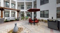 This thoughtfully designed courtyard is nestled within a contemporary multi-story building, creating an enclosed, semi-private outdoor retreat. The surrounding architecture features a crisp white façade with large, dark-framed windows and latticed balconies that overlook the space, lending a sense of verticality and enclosure while maintaining an open-air atmosphere.

The courtyard's flooring combines herringbone-patterned brick pavers with large-format stone tiles, creating a visually dynamic surface that grounds the various functional zones. At the heart of the outdoor kitchen area, a stainless steel built-in grill sits atop a dark stone counter, providing a practical amenity for outdoor entertaining. Nearby, a bold dark wood pergola anchors a comfortable lounge area furnished with cushioned seating and a low table, offering a shaded retreat.

A round dining table with bold red cushioned chairs and a matching market umbrella provides an additional gathering spot suited for al fresco meals. Decorative landscaping elements, including river rock beds, ceramic vase accents, small ornamental trees, and low plantings, add texture and visual interest throughout. The overall ambiance is modern and inviting, balancing functional amenities with clean, contemporary aesthetics.