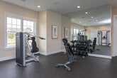 Located at 700 SR-96 E in Bonaire, Georgia, this single-floor fitness center is thoughtfully designed to support a comprehensive workout experience. The space features durable rubber flooring throughout, providing safety and comfort during exercise sessions.

The cardio zone is well-appointed with multiple treadmills, elliptical trainers, and stationary bikes, each equipped with individual display screens. Large multi-pane windows line the walls, flooding the area with natural light and offering views of the surrounding neighborhood and outdoor landscape.

The strength training section includes a cable crossover machine, a selection of selectorized weight machines for targeted muscle training, and a full dumbbell rack with a wide range of free weights. A wall-length mirror in the free weights area enhances the sense of space and allows users to monitor their form during workouts.

Recessed lighting is installed throughout the ceiling, ensuring consistent illumination across all areas of the gym. A wall-mounted television is positioned in the strength training zone for entertainment. The warm, neutral-toned walls contribute to an inviting atmosphere, while in-ceiling speakers suggest built-in audio capabilities. The space is well-ventilated with visible HVAC vents and maintains a clean, organized layout suited for a community fitness facility.