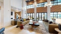 Located at 2022 N Beckley Ave in Dallas, TX, this sophisticated single-floor communal lounge and gathering space makes a striking first impression with its dramatic double-height ceilings and bold exposed brick accent walls. The expansive open floor plan is anchored by a generously sized living area furnished with plush, earth-toned sofas, tufted ottomans, and layered area rugs, all illuminated by sculptural globe pendant chandeliers that add a modern, artistic touch. Natural light pours in abundantly through tall, black-framed floor-to-ceiling windows and folding glass doors, which open directly onto an outdoor patio furnished with vibrant yellow seating and shaded umbrellas. A casual dining nook features rich leather banquette seating, marble-topped tables, and chevron-patterned hardwood flooring, creating an inviting space for conversation and community. Lush hanging greenery cascades from elevated planters, softening the industrial elements and adding life to the interior. Built-in shelving and a coffee bar area with warm wood cabinetry provide functional amenities, while a separate lounge nook with navy upholstered chairs offers a quieter retreat. The overall ambiance masterfully blends industrial character with refined, contemporary comfort.