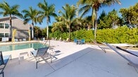 Located in Estero, Florida, this resort-style outdoor amenity area offers a quintessential Southwest Florida living experience. At its centerpiece is a generously sized rectangular swimming pool with crystal-clear blue water, flanked on all sides by a broad, light-colored concrete pool deck designed to accommodate ample sunbathing space. Rows of modern lounge chairs and chaise lounges are thoughtfully arranged around the pool, providing residents with plenty of options for relaxation in the abundant Florida sunshine. Elegant pergola-style cabana structures with yellow drapery accents add a touch of resort luxury and provide shaded retreats along the poolside. The area is beautifully framed by a dense canopy of tall royal palm trees and lush tropical greenery, creating a private, serene atmosphere. A covered colonnade with stone-clad columns and ceiling fans borders one side of the pool deck, offering a shaded transition zone between the interior clubhouse and the outdoor area. A dedicated restroom facility, finished with a terracotta-tiled roof and natural stone detailing, is conveniently accessible from the pool deck. A freestanding hammock adds a relaxed, leisure-focused touch to the space, completing the tropical ambiance of this well-appointed outdoor amenity.