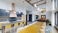 This thoughtfully designed single-floor space offers a seamless open-plan layout that blends functionality with contemporary style. The kitchen area is a standout feature, boasting a professional-grade range, a bold navy blue tile backsplash, and light wood cabinetry paired with a sleek ventilation hood. Pendant lighting in warm metallic tones illuminates the workspace, adding both task lighting and visual interest. A long, natural wood communal dining table anchors the center of the space, surrounded by an ample number of cream and yellow accent chairs that inject a vibrant pop of color into the palette. The soaring ceilings are accented by exposed wood beam detailing finished in gold tones, lending warmth and architectural character to the otherwise crisp white interior. Adjacent to the dining area, a comfortable lounge zone features rich cognac leather seating arranged around a mounted flat-screen display, creating an inviting setting for relaxation or casual gatherings. The open sightlines between the kitchen, dining, and lounge areas foster a sense of connectivity and spaciousness throughout. Neutral tile flooring, recessed lighting, and carefully chosen pendant fixtures tie the entire space together into a cohesive, modern aesthetic well-suited for communal use.
