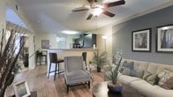 This single-floor residence offers a thoughtfully designed open-concept layout that integrates the living, dining, and kitchen areas into one cohesive and functional space. The living area is warm and inviting, anchored by rich hardwood-style flooring that extends throughout the main living spaces, lending a sense of warmth and continuity. A ceiling fan with a light fixture is centrally positioned overhead, providing both comfort and ambient illumination.

The kitchen is defined by dark cabinetry with modern hardware, stainless steel appliances, and a breakfast bar with elevated seating — a practical feature that doubles as a casual dining area. The bar-height counter creates a natural visual boundary between the kitchen and the living space without sacrificing the open feel of the floor plan.

The living area is bright and well-lit, benefiting from a combination of natural and artificial light sources, including a tall floor lamp that adds a contemporary touch. Neutral tones on the walls complement the overall design palette, creating a calm and balanced ambiance. The space is versatile and welcoming, ideal for both relaxed everyday living and entertaining guests.