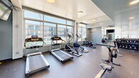 This fitness center offers a comprehensive workout environment designed to support a wide range of exercise routines. The single-floor space is framed by large, floor-to-ceiling windows along multiple walls, flooding the interior with abundant natural light and providing open views of the surrounding outdoor area. The durable rubber flooring throughout the exercise room provides both safety and functionality for a variety of workouts.

The cardio zone is well-appointed with multiple treadmills, elliptical machines, and stationary bikes lined up facing the expansive windows. A dedicated strength training section features a full rack of dumbbells in graduated weights, a cable weight machine, and additional resistance equipment, complemented by wall-to-wall mirrors that enhance the sense of space and allow for proper form monitoring. A mounted flat-screen television and a water fountain add to the convenience of the space.

A sliding glass door opens directly to an outdoor turf training area, equipped with battle ropes and agility equipment, offering an al fresco workout option. The glass-fronted entrance provides a bright, welcoming entry point. Flush-mounted ceiling lights ensure consistent illumination throughout. Overall, the fitness center delivers a clean, modern, and motivating atmosphere for residents seeking a complete and versatile exercise experience.