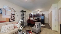 This single-floor residence showcases an open-concept layout that connects the living, dining, and kitchen areas into one cohesive and functional space. The living area is comfortably sized, featuring neutral-toned walls and a mix of flooring that transitions between carpet and hard surface, creating defined zones within the open plan. A stylish arc floor lamp and a decorative display shelf with glass panels add visual interest alongside tasteful décor, including floral arrangements and accent artwork.

The dining area sits centrally within the space, accommodating a dark-toned table with upholstered chairs, illuminated by a chandelier overhead that adds a touch of elegance. The kitchen, seamlessly integrated into the layout, features rich dark cabinetry, black appliances including a refrigerator and range, and what appears to be a granite-style countertop that extends to form a breakfast bar. Recessed and pendant lighting throughout ensures the space is well-lit. The overall ambiance is warm and inviting, with a cohesive color palette of deep browns, reds, and neutral tones that lend the space a polished, contemporary feel.