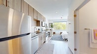 This thoughtfully designed studio offers a seamless open-concept layout that maximizes every inch of space. The kitchen runs along one wall and is outfitted with stainless steel appliances — including a full-size range, over-the-range microwave, dishwasher, and refrigerator — complemented by light wood cabinetry with brushed gold hardware and a classic white subway tile backsplash. A stainless steel sink and generous counter space complete the functional yet stylish cooking area. Light wood-tone flooring flows continuously throughout, connecting the kitchen to the bright and airy sleeping area. A large picture window dominates the far wall, flooding the space with natural light and framing lush tree canopy views that bring the outdoors in. The sleeping area is open and well-proportioned, offering ample room for a full-size bed and additional furnishings. The bathroom features marble-style tile flooring, a light wood vanity with brushed gold hardware, a large mirror, and crisp white walls — creating a clean, spa-like feel. Recessed lighting throughout the unit ensures a consistently bright atmosphere. The overall aesthetic is modern and minimal, with warm natural wood tones, white walls, and gold accents creating a cohesive and inviting living environment.