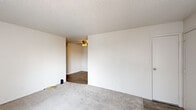 This efficient single-floor unit is thoughtfully laid out to maximize every square foot, featuring one bedroom, one bathroom, a living room, and a kitchen. The living room is a bright, open space with neutral carpet flooring, white walls, a textured ceiling, and a wall-mounted HVAC unit. Two windows fitted with vertical blinds allow natural light to filter in, and a ceiling fan in the adjacent area adds to the overall comfort of the space. The bedroom continues the carpeted flooring and clean white walls, featuring a window with vertical blinds that brings in natural light, creating a calm and functional retreat. The galley-style kitchen is well-appointed with white cabinetry lining both sides, light stone-patterned countertops, a stainless steel sink, a white refrigerator, a range with electric cooktop, and wood-look vinyl flooring. Track lighting overhead brightens the workspace, and a window at the end of the kitchen adds ventilation and light. The bathroom is clean and straightforward, featuring wood-look vinyl flooring, a toilet, and a bathtub. Throughout the unit, a consistent palette of white walls and light flooring creates a cohesive, easy-to-maintain living environment.