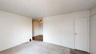 This efficient single-floor unit is thoughtfully laid out to maximize every square foot, featuring one bedroom, one bathroom, a living room, and a kitchen. The living room is a bright, open space with neutral carpet flooring, white walls, a textured ceiling, and a wall-mounted HVAC unit. Two windows fitted with vertical blinds allow natural light to filter in, and a ceiling fan in the adjacent area adds to the overall comfort of the space. The bedroom continues the carpeted flooring and clean white walls, featuring a window with vertical blinds that brings in natural light, creating a calm and functional retreat. The galley-style kitchen is well-appointed with white cabinetry lining both sides, light stone-patterned countertops, a stainless steel sink, a white refrigerator, a range with electric cooktop, and wood-look vinyl flooring. Track lighting overhead brightens the workspace, and a window at the end of the kitchen adds ventilation and light. The bathroom is clean and straightforward, featuring wood-look vinyl flooring, a toilet, and a bathtub. Throughout the unit, a consistent palette of white walls and light flooring creates a cohesive, easy-to-maintain living environment.