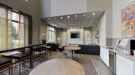 This single-floor communal space is thoughtfully designed to serve as a versatile social and lounge area. The layout is open and spacious, anchored by dramatically high ceilings that create an airy, expansive atmosphere. Recessed lighting throughout ensures the space is well-illuminated, complemented by generous natural light streaming through large floor-to-ceiling windows dressed with sheer golden-hued drapes, offering views of a landscaped outdoor pool area.

The lounge is divided into distinct functional zones. A comfortable seating area features a large sectional sofa in charcoal gray, paired with distinctive natural wood burl coffee tables and a wall-mounted flat-screen television. Surrounding round dining tables with wire accent chairs and a dark bar-height communal table with sleek stools provide additional gathering options.

A dedicated kitchenette area is outfitted with clean white cabinetry, a light gray tile backsplash, stainless steel appliances including a built-in microwave and under-counter refrigerator, and a coffee station. Bold, colorful abstract artwork displayed above the counter adds a vibrant focal point to this area.

The flooring consists of patterned carpet throughout, contributing warmth and texture. Large-scale abstract and monochromatic artwork decorates the walls, reinforcing the space's contemporary, curated aesthetic.