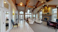 This single-story clubhouse is defined by its striking architectural character, centered around soaring vaulted ceilings adorned with prominent exposed wood beam trusses that lend a warm, lodge-inspired aesthetic to the open interior. The main gathering area is generously proportioned, seamlessly blending a tiled entry zone with a carpeted lounge space furnished with comfortable seating arranged around a central coffee table. A standout feature of the space is the full brick fireplace with a wood mantel, framed by tall multi-pane windows that flood the interior with natural light and offer views of the surrounding outdoor areas, including a deck and pool. Track lighting and ceiling fans complement the natural illumination throughout. A dining area near the entry accommodates a formal table and chairs, positioned to take full advantage of the expansive window walls and exterior access doors. Toward the rear of the clubhouse, a functional kitchen is equipped with standard appliances, oak cabinetry, and tile flooring, suitable for event preparation. Additional doors provide access to restrooms and utility areas. The overall ambiance is open, welcoming, and well-suited for community gatherings and social events.