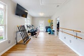 This single-floor fitness center is thoughtfully designed to accommodate a wide range of workout styles across multiple distinct zones. The space features warm light hardwood flooring throughout, neutral-toned walls, and textured ceilings that create a clean, inviting atmosphere.

The main cardio room is generously proportioned, offering ample space for multiple treadmills, an elliptical, and a stationary bike. Large windows line the walls, flooding the room with natural light and providing outdoor views. A wall-mounted flat-screen television enhances the workout experience, and a multi-station strength machine and dumbbell rack round out the equipment offerings.

An adjoining studio room serves as a dedicated space for group fitness and low-impact training. It features a wall-mounted ballet barre, suspension training straps, and smooth open floor space ideal for stretching, yoga, or aerobics. A glass-paned door provides an additional entry point and adds to the bright, airy feel of the room.

A third area connects the zones and houses an array of fitness accessories, including cycling bikes, a rack of weighted medicine balls, stacked exercise mats, and colorful resistance equipment. This section also benefits from a glass-paned exterior door that opens to the outdoors.

Together, the interconnected rooms form a comprehensive, well-organized fitness facility suited for both individual and group exercise activities.