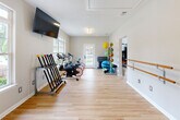 This single-floor fitness center is thoughtfully designed to accommodate a wide range of workout styles across multiple distinct zones. The space features warm light hardwood flooring throughout, neutral-toned walls, and textured ceilings that create a clean, inviting atmosphere.

The main cardio room is generously proportioned, offering ample space for multiple treadmills, an elliptical, and a stationary bike. Large windows line the walls, flooding the room with natural light and providing outdoor views. A wall-mounted flat-screen television enhances the workout experience, and a multi-station strength machine and dumbbell rack round out the equipment offerings.

An adjoining studio room serves as a dedicated space for group fitness and low-impact training. It features a wall-mounted ballet barre, suspension training straps, and smooth open floor space ideal for stretching, yoga, or aerobics. A glass-paned door provides an additional entry point and adds to the bright, airy feel of the room.

A third area connects the zones and houses an array of fitness accessories, including cycling bikes, a rack of weighted medicine balls, stacked exercise mats, and colorful resistance equipment. This section also benefits from a glass-paned exterior door that opens to the outdoors.

Together, the interconnected rooms form a comprehensive, well-organized fitness facility suited for both individual and group exercise activities.