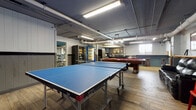 This single-floor recreational space is thoughtfully designed to serve as a social and leisure hub. The main game room is the centerpiece, featuring a full-size Butterfly ping-pong table and a red-felted billiards table, both set atop durable wood-look flooring. Exposed pipes and industrial-style fluorescent lighting run across the ceiling, lending the space a casual, urban character. A wall-mounted flat-screen television and wood-paneled wainscoting add to the relaxed atmosphere.

Adjacent to the game area is a well-appointed kitchen outfitted with teal cabinetry, dark stone countertops, stainless steel appliances including a full-size refrigerator and dishwasher, a microwave, and a deep sink. A kitchen island with bar-height stools provides an informal dining and gathering spot. A small window allows natural light to filter into the kitchen area.

A dedicated lounge corner features a large black leather sofa positioned beneath a rustic moose-themed wall art piece, creating a cozy retreat within the larger space. Beverage and snack vending machines are conveniently located near a mirrored wall, with an adjacent fitness area visible in the reflection. The overall ambiance is casual, functional, and well-suited for communal recreation and relaxation.