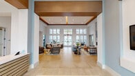 This single-floor amenity center showcases a thoughtfully designed coastal aesthetic throughout its various interconnected spaces. The grand entrance greets visitors with soaring double-height ceilings, a coffered wood-accented ceiling detail, and large floor-to-ceiling glass doors that flood the space with natural light. A sleek reception desk, clad in light wood slat paneling and topped with white quartz, anchors the arrival experience, accented by vibrant ocean-inspired artwork.

The expansive main lounge features warm tile flooring, multiple seating arrangements with woven and upholstered furnishings, and access to adjacent rooms. A bright café-style dining area offers abundant window views to the outdoors, furnished with round tables and contemporary chairs in a soft blue-and-white palette.

The clubroom stands out with a long white quartz island, bar-height seating, light wood cabinetry, dual wall-mounted televisions, and striking teal blown-glass pendant lights. Decorative surfboards add a playful coastal touch to the walls.

The fitness center is divided into distinct zones: a cardio and cycling studio with floor-to-ceiling mirrors, wall-mounted screens, and large windows overlooking the pool, alongside a separate strength training room equipped with professional machines on colorful tile-patterned flooring. A quiet reading nook with large windows completes this well-appointed facility.