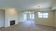 This single-story home showcases an open-concept living area defined by thoughtful architectural details. A standout feature is the elegant arched entryway flanked by decorative white columns with classic pedestal bases, which elegantly divides the living and dining spaces while maintaining an open, connected feel. The living room is anchored by a built-in fireplace with a clean white surround, adding both warmth and visual interest to the space. Plush wall-to-wall carpeting in a neutral beige tone runs throughout, contributing to the overall comfort and cohesion of the interior. The walls are finished in a soft, warm neutral palette that complements the light-colored trim and baseboards. Natural light flows generously into the space through multiple windows, illuminating the rooms with a bright, airy quality. A pendant light fixture with a classic dome shade hangs above the dining area, providing focused lighting and a subtle decorative accent. A ceiling fan is also visible in the adjoining room, enhancing both comfort and functionality. The overall ambiance is calm, neutral, and move-in ready, offering a versatile canvas for a variety of interior design styles.