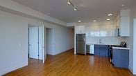 This single-floor unit features a contemporary open-plan design that seamlessly integrates the living and kitchen areas. The kitchen is equipped with stainless steel appliances, including a full-sized refrigerator and dishwasher, complemented by clean white cabinetry that extends to the ceiling, providing generous storage. A stainless steel sink and modern fixtures complete the functional kitchen setup. The flooring throughout is warm-toned hardwood, adding a welcoming contrast to the crisp white walls. Recessed lighting and a track lighting system in the main living area ensure consistent, well-distributed illumination. A hallway connects to additional rooms, featuring louvered bifold doors that hint at in-unit storage or laundry access. The bedroom area benefits from a large window that draws in natural light, contributing to an airy atmosphere. Glass-paneled interior doors enhance the sense of openness between spaces. The overall design is clean and minimalist, with neutral tones, modern finishes, and a practical layout that maximizes the use of available space throughout the unit.