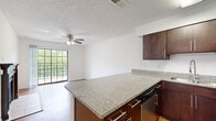 This thoughtfully designed one-bedroom, one-bathroom unit offers a compact yet functional layout across a single floor, making efficient use of every square foot. The open-concept kitchen anchors the living space with rich, dark-stained cabinetry, granite countertops extending into a convenient peninsula, stainless steel appliances including a range, microwave, and refrigerator, and a built-in sink. Wood-look flooring flows throughout the main living areas, lending warmth and visual continuity. The living room is a standout, featuring a classic wood-mantled fireplace with tile surround and a sliding glass door that opens to a private balcony overlooking lush greenery, filling the space with natural light. A ceiling fan completes the comfortable atmosphere. The bedroom is a well-sized retreat with wood-look flooring, a ceiling fan, and a window with blinds providing natural light. The bathroom features a full tub-shower combination with clean white tile accented by a decorative mosaic border. A dedicated laundry area houses a full-size washer and dryer beneath dark wood cabinetry, adding everyday convenience. Additional storage is available via a hallway closet, rounding out this efficiently organized unit.