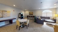 This single-floor recreational lounge is thoughtfully designed to serve as a versatile social hub, offering a range of entertainment options across its open, interconnected spaces. The interior features neutral-toned walls, recessed lighting, and plush carpeting throughout, creating a welcoming and comfortable atmosphere.

The main game area is well-equipped with standout amenities, including an arcade machine, a full-size shuffleboard table, and a giant Connect Four set, providing entertainment options for a variety of interests. A round dining table surrounded by woven accent chairs offers a casual spot for gathering, while a sleek wall-mounted counter with bar-height stools serves as a high-top station, accented by colorful artwork above.

A generously sized sectional sofa anchors a lounge area positioned near an arched window that allows natural light to filter in. Additional seating, including upholstered armchairs and ottomans, is distributed throughout the space, accommodating both quiet relaxation and group socializing.

A transitional hallway area features a plush yellow armchair with ottoman, framed artwork, and a decorative indoor tree, adding warmth and a residential feel. The overall design balances playful functionality with a polished, approachable aesthetic.
