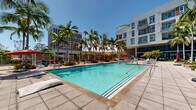 Located at 5075 Camino de la Siesta in San Diego's Mission Valley neighborhood, this outdoor amenity space showcases a generously sized resort-style swimming pool as its centerpiece. The pool features crystal-clear turquoise water and is framed by a wide, tiled pool deck designed to accommodate an abundance of lounge chairs, chaise lounges, and shade umbrellas in bold red and blue tones. Towering palm trees are planted throughout the courtyard, providing a tropical atmosphere and natural shade under the abundant Southern California sunshine. Structured shade cabanas with slatted wooden screens line the perimeter, offering semi-private seating areas ideal for relaxing in comfort. Lush tropical plantings and decorative landscaping add greenery and visual depth throughout the space. The courtyard is embraced on multiple sides by a modern, multi-story residential building featuring contemporary architecture with balconies, warm wood-toned paneling, and a neutral stucco facade. An outdoor grilling and entertaining station is also visible, further enhancing the community-focused amenity offerings. The overall ambiance is vibrant and resort-like, perfectly suited to San Diego's year-round temperate climate and outdoor lifestyle.