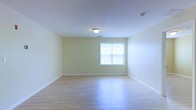 This single-floor space presents a clean, straightforward layout with a welcoming and functional design. The room is finished with light hardwood flooring that runs throughout, lending warmth and a sense of openness to the interior. The walls are painted in a soft, pale yellow tone, creating a neutral and inviting atmosphere that pairs well with the light tones of the floor.

A centrally positioned window on the far wall allows natural light to filter into the room, brightening the space and highlighting the reflective quality of the flooring below. The window is framed simply, consistent with the understated aesthetic of the room overall.

The ceiling is smooth and white, fitted with a single flush-mount light fixture that provides ambient overhead lighting. A smoke or carbon monoxide detector is also visible in the upper corner, noting the space's attention to safety features. White baseboards run along the perimeter of the room, offering a crisp contrast to the yellow walls and wood flooring.

The room's layout is open and unobstructed, making it adaptable for a variety of uses. The overall ambiance is modest, bright, and move-in ready, suited for practical, everyday living.