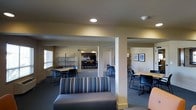 This single-floor space functions as a multi-purpose communal area, thoughtfully organized into distinct zones that cater to a variety of activities. The layout is defined by structural columns and open passageways that divide the space into functional areas while maintaining an open, connected feel throughout.

The main lounge area is anchored by a striped upholstered sofa and additional seating, providing a comfortable spot for relaxation or informal gatherings. Adjacent to this are dedicated work and study zones equipped with tables and task chairs, ideal for individual or group use. Metal shelving units line portions of the walls, offering practical storage or display space.

Toward the rear of the space, a kitchen area is visible, outfitted with dark cabinetry, appliances, and a dining table with chairs — supporting meal preparation and communal dining. Additional seating and work table arrangements are positioned throughout, enhancing the space's versatility.

Recessed and overhead lighting illuminates the interiors, complemented by natural light filtering through window blinds. Framed artwork on the walls adds a modest decorative touch. The neutral color palette, comprising beige walls and gray carpeting, creates a calm, functional ambiance throughout.