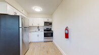 This single-floor unit is laid out with a practical, open-feeling floor plan that connects its key living spaces efficiently. The kitchen is a standout area, outfitted with white shaker-style cabinetry featuring matte black hardware, light stone-toned countertops, a subway tile backsplash, and stainless steel appliances including an electric range with over-the-range microwave. Light wood-look flooring runs through the kitchen and entry areas, contributing to a clean, contemporary aesthetic.

The living area transitions from hard flooring to neutral-toned carpet, offering a comfortable feel underfoot. A front-facing window allows natural light to fill the space, with views of the surrounding neighborhood visible through slatted blinds. The entry door is positioned conveniently off this main area.

The bedroom is generously proportioned, featuring the same neutral carpet and walls in a warm greige tone. It includes both a closet and an additional door, providing practical storage and access options. A window brings in natural light.

The bathroom is finished with large-format tile flooring in a warm beige wood-look pattern, bright white walls with textured tile accents, and a natural wood-toned vanity counter, creating a clean and modern feel. Overall, the unit maintains a consistent, understated design throughout.