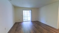 This single-floor space is defined by its clean, straightforward layout and warm, inviting finishes. The main living area showcases rich, dark hardwood flooring that runs throughout, lending a sense of warmth and continuity to the room. The walls are finished in a soft, neutral cream tone that complements the flooring and contributes to a bright, open atmosphere.

A standout feature of the space is the sliding glass door, which serves as both a functional entry point to the outdoors and a primary source of natural light. The door is framed by vertical blinds, offering adjustable privacy and light control, while the view beyond reveals lush, mature greenery. This connection to the outdoors enhances the overall sense of openness within the room.

The ceiling features a textured finish and is kept at a standard height, contributing to the room's comfortable proportions. The smooth, plain walls provide a versatile backdrop, making the space adaptable to a variety of interior styles and furnishing arrangements. Overall, the space offers a practical and well-lit environment with durable, attractive finishes throughout.